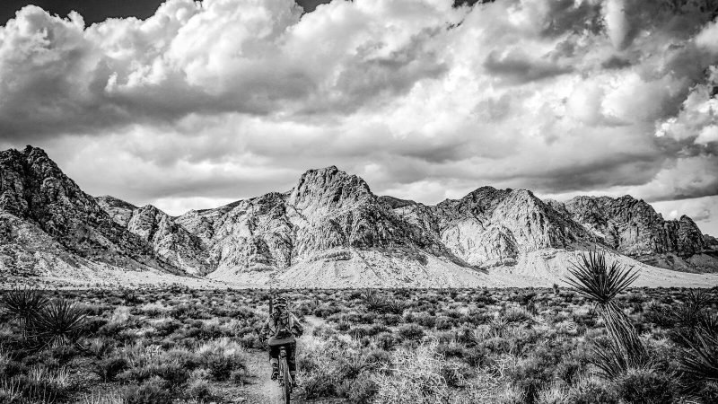 A lone cyclist rides through a desert landscape towards mountains.